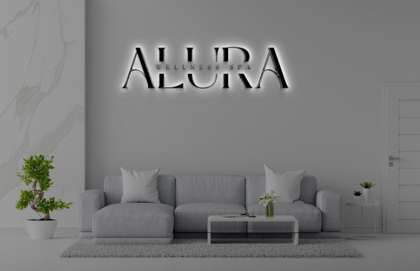 Backlit 'ALURA Wellness Spa' logo above light gray sectional sofa, marble accent wall, bonsai, glass coffee table.