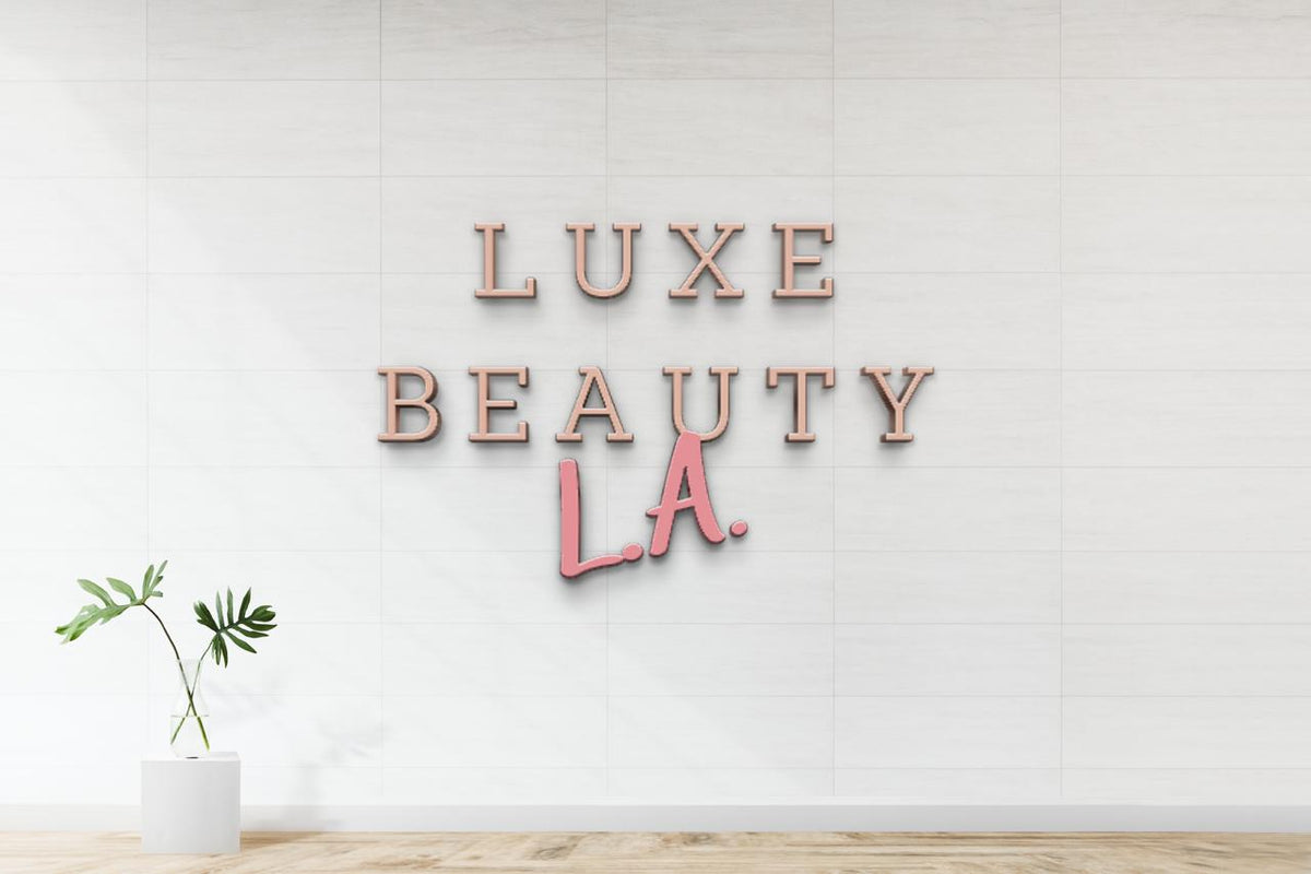 Rose-gold and pink 3D letters reading 'LUXE BEAUTY LA.' on white tiled wall above light wood floor, potted plant.