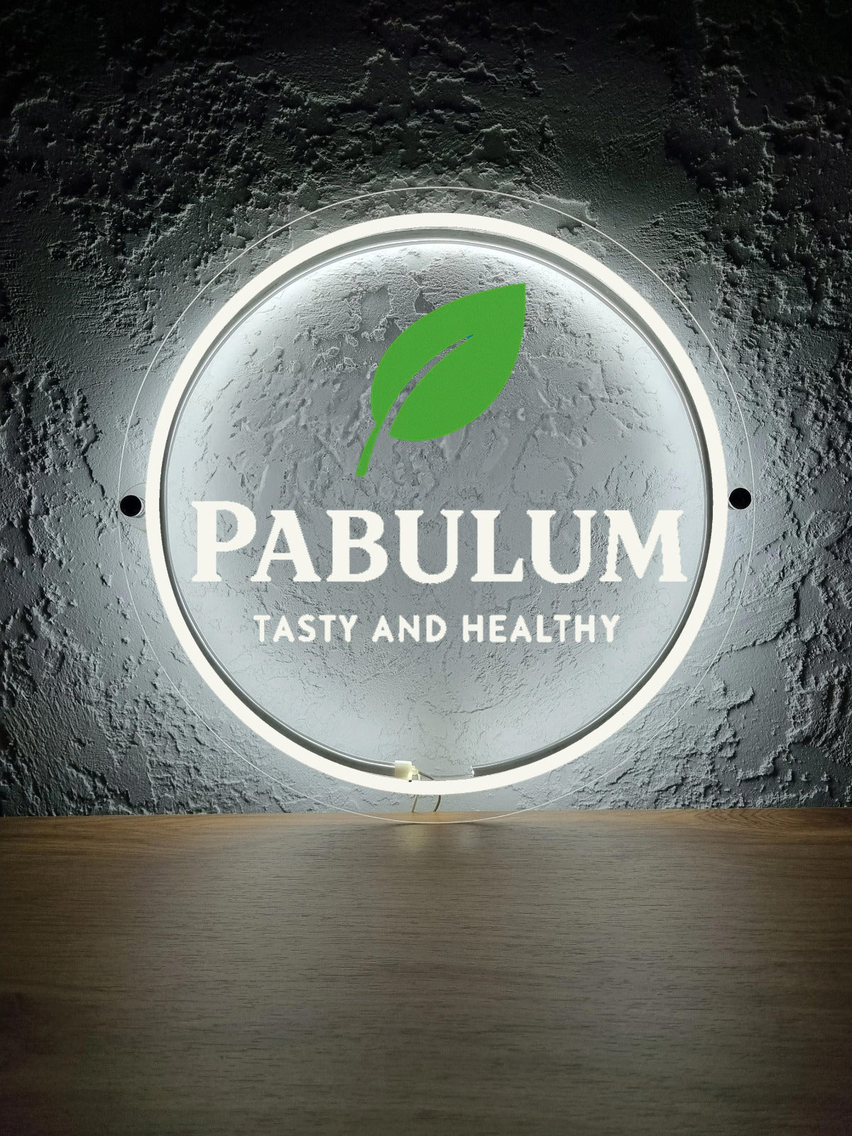Circular illuminated sign with green leaf logo, bold PABULUM text and 'TASTY AND HEALTHY' slogan on textured gray wall.