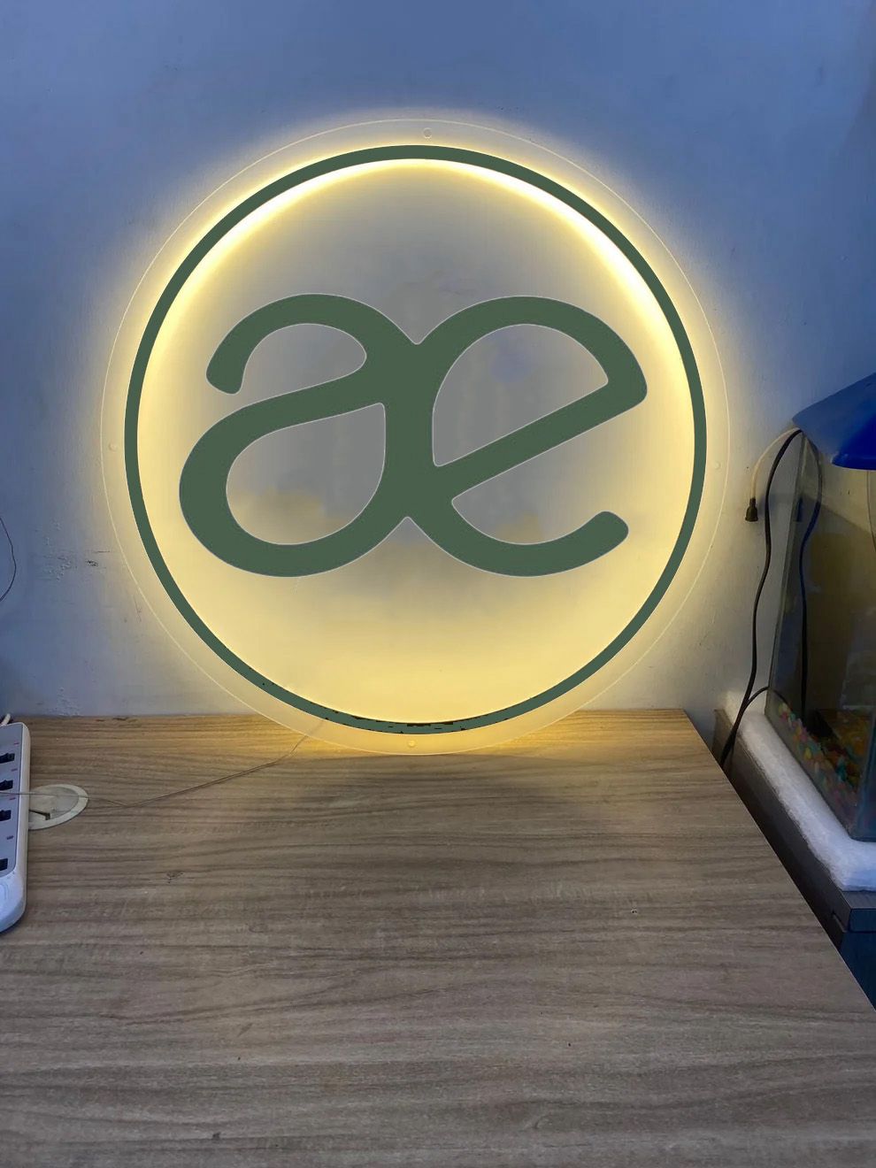 LED backlit round sign with green ae logo and thin circle on wooden desk, warm yellow halo, aquarium nearby.