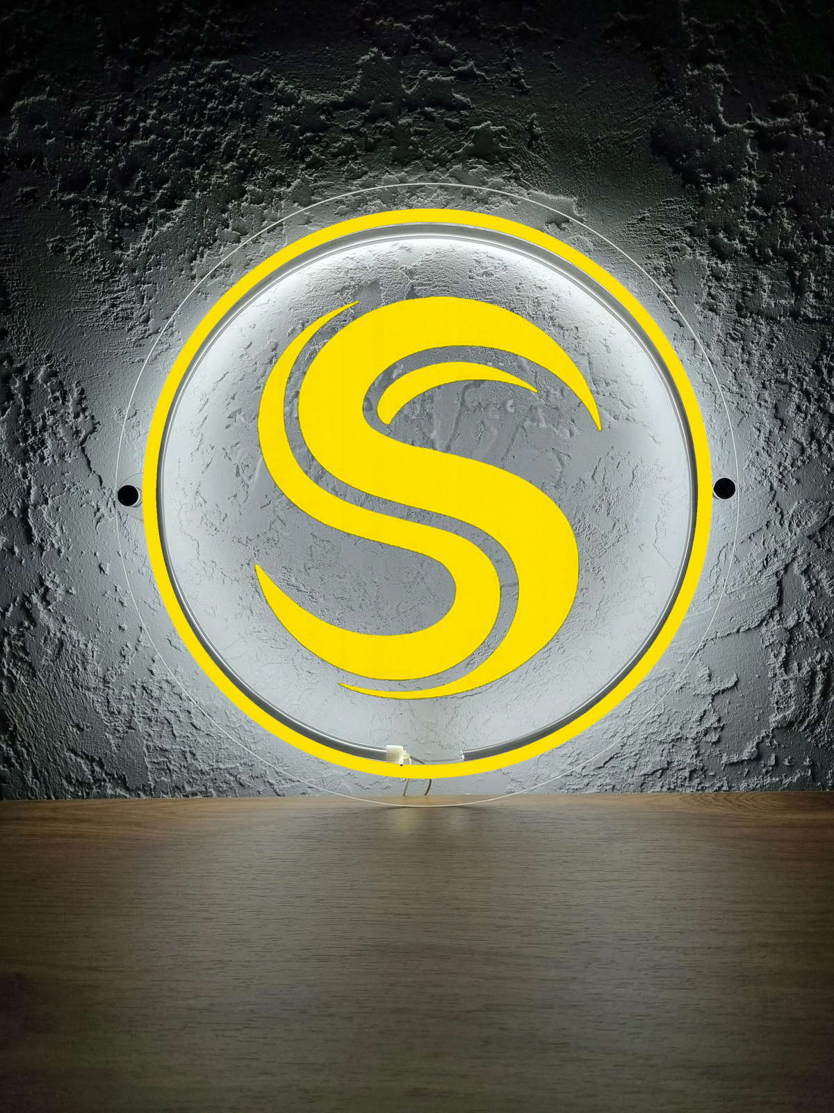 Round illuminated sign with yellow stylized 'S' and neon ring mounted on textured gray concrete wall above wood.