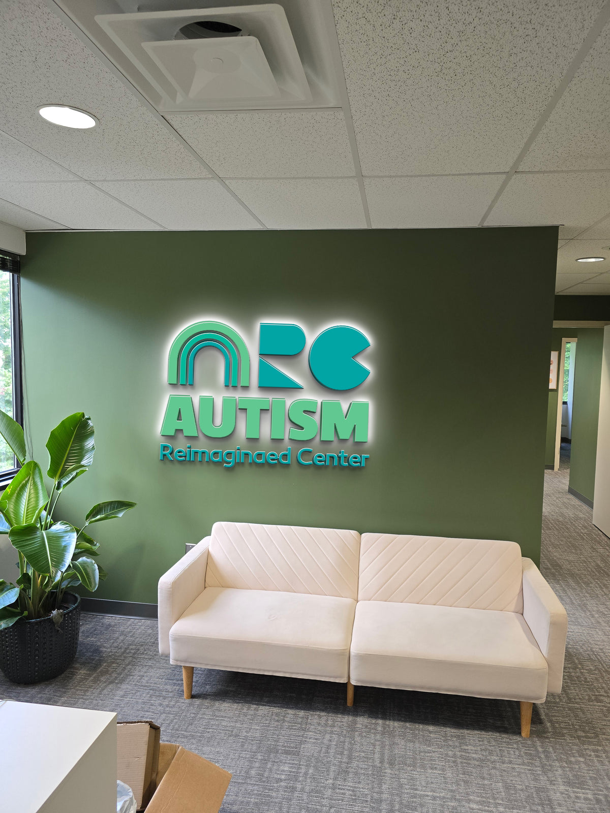 Green reception wall with illuminated teal AUTISM Reimaginaed Center logo, cream sofa, potted plant, gray carpet.