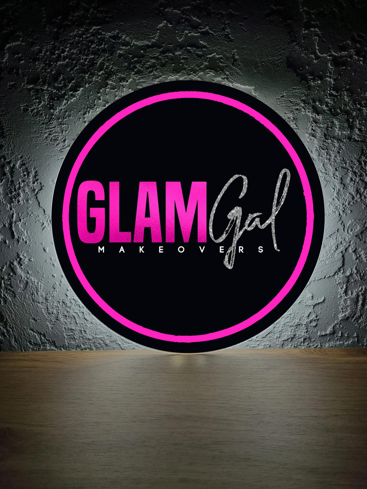 Round black sign with neon pink ring and bold GLAM plus script Gal Makeovers on textured gray wall above wood floor.