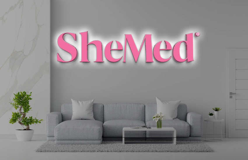 Minimalist gray living room with light-gray sofa, glass coffee table, potted plants and illuminated pink 'SheMed' logo.