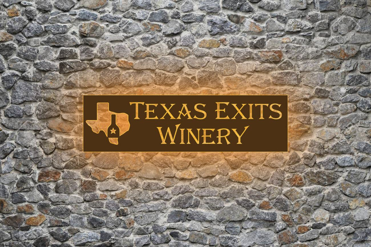 Brown sign 'Texas Exits Winery' with gold Texas outline and wine bottle emblem on gray stone wall with amber glow.