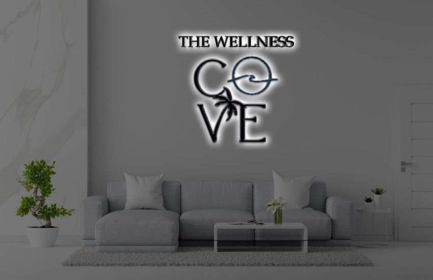 Gray living room with sofa, coffee table, potted plants, illuminated wall logo 'THE WELLNESS COVE' with wave and palm.