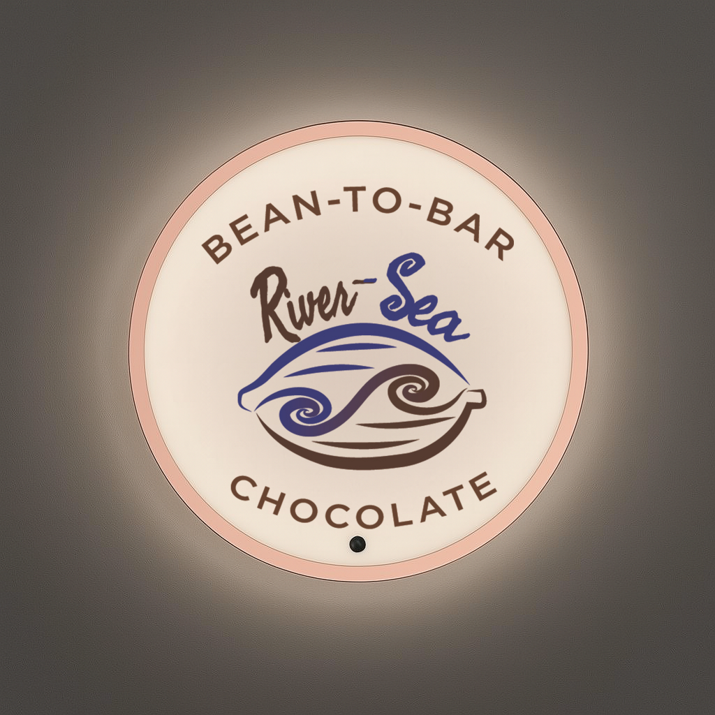 Illuminated round sign with pink rim reading 'Bean-to-Bar Chocolate' and 'River-Sea' over blue-brown cocoa pod logo.