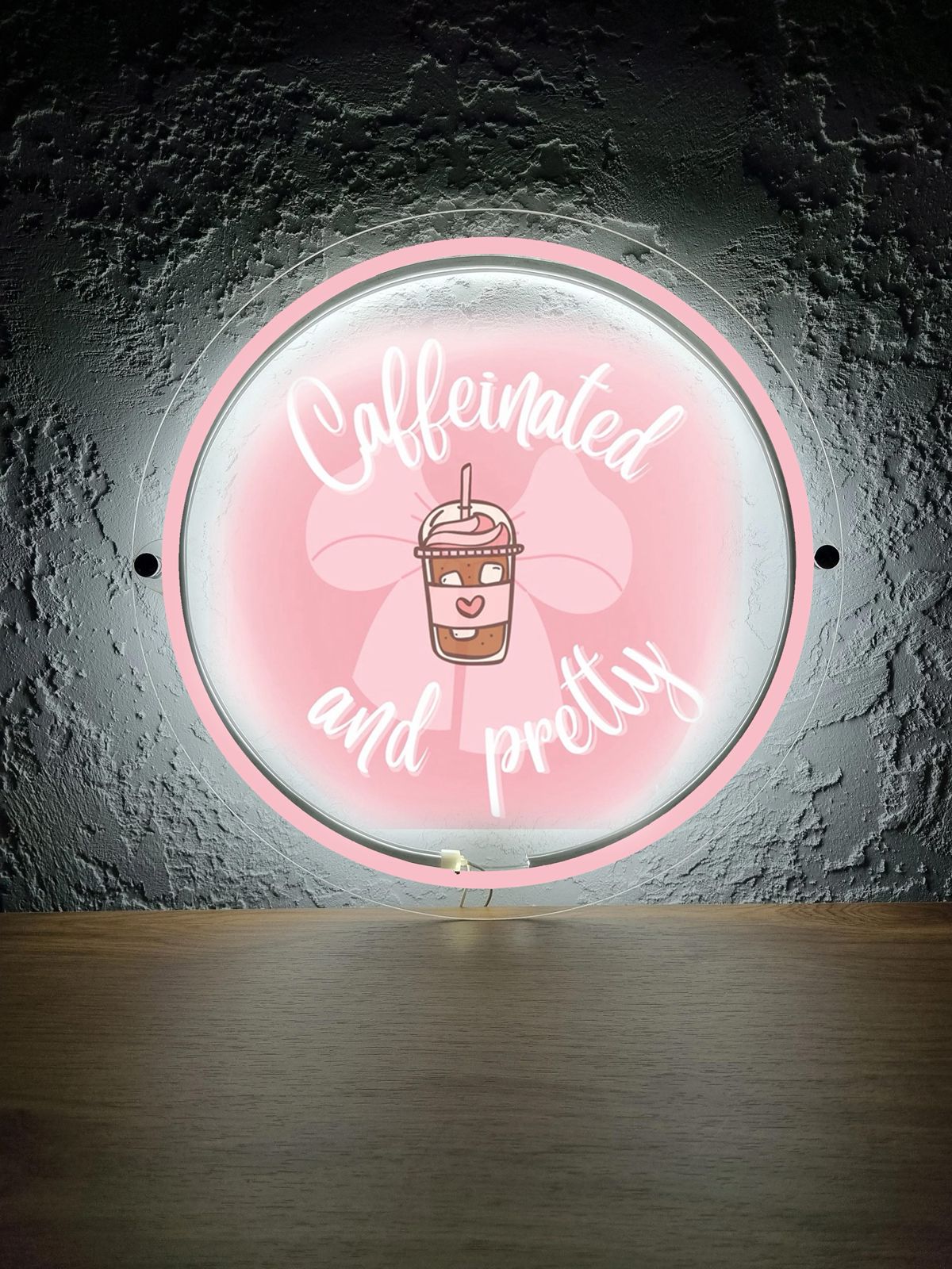 Round pink neon wall sign reading 'Caffeinated and pretty' with cartoon iced coffee cup over wood shelf.