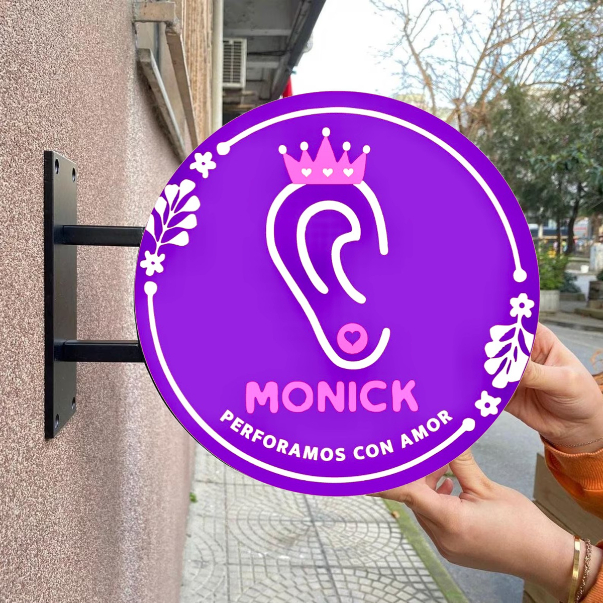 Hands holding round purple sign with white ear logo and pink crown, MONICK and PERFORAMOS CON AMOR on pink wall.