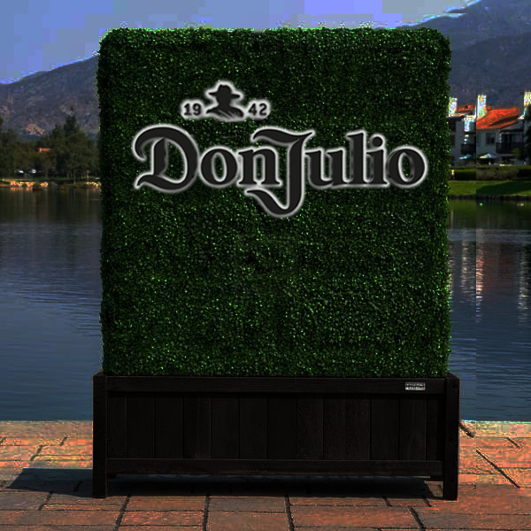 A square green hedge wall with a large Don Julio 1942 logo mounted on a dark wooden planter by a lakeside.