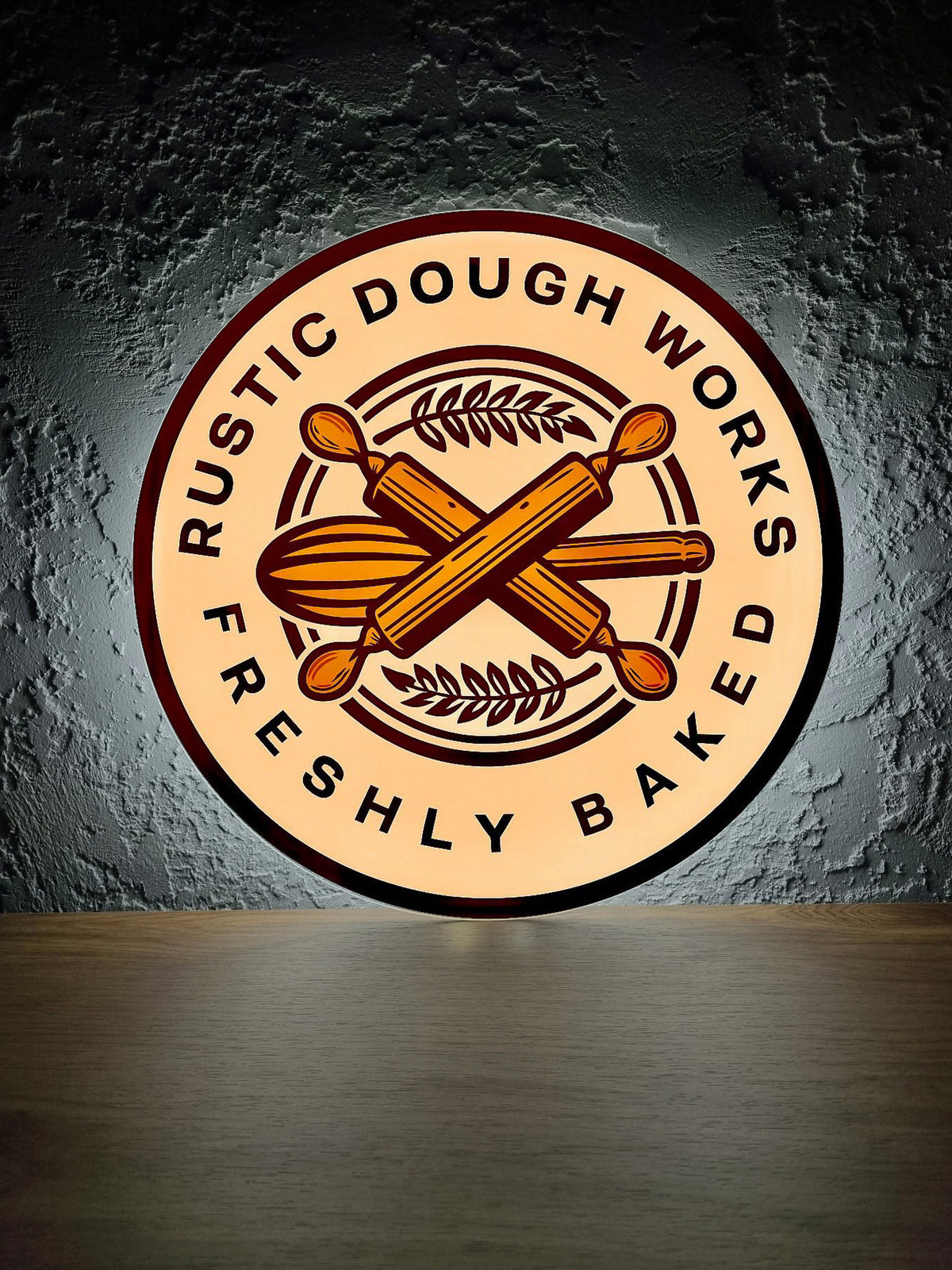 Beige Rustic Dough Works bakery logo with maroon rim, crossed rolling pins and wheat on gray textured wall.