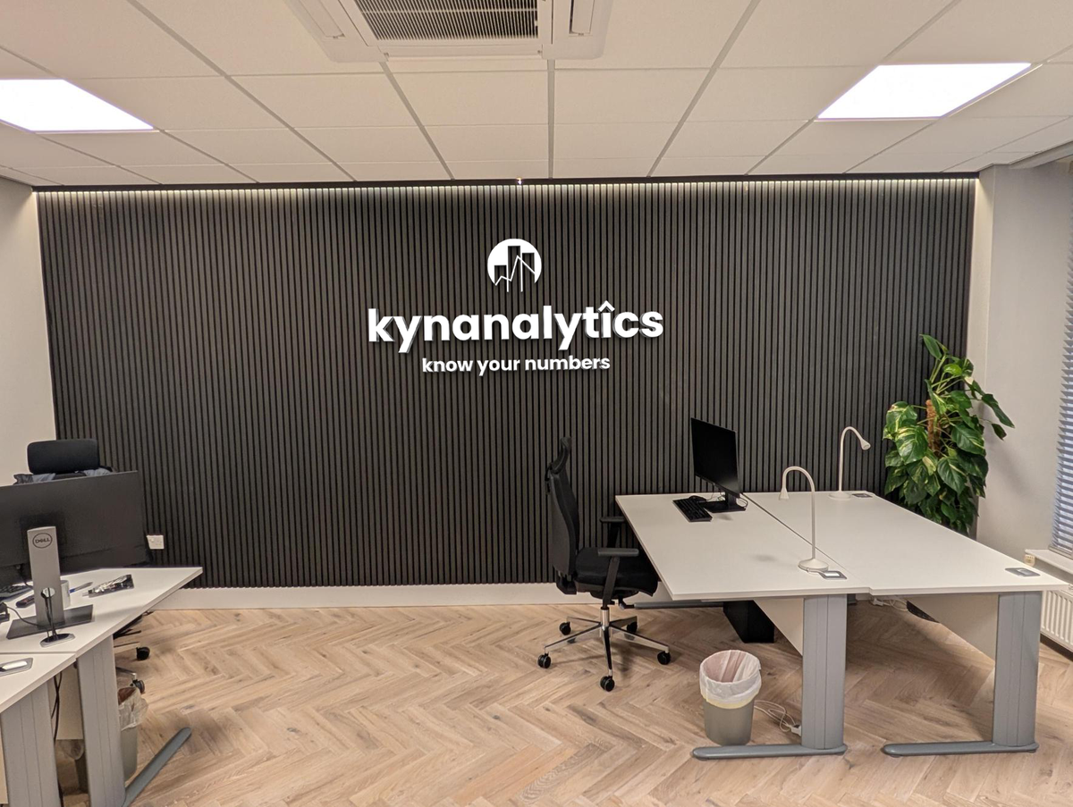 Office with black vertical slatted wall backlit by white 'kynanalytics' logo and 'know your numbers' tagline, white d.