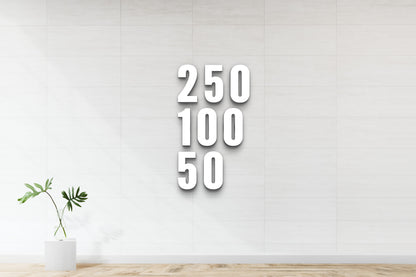 Large white 3D numbers 250, 100, 50 stacked vertically on light tiled wall, green monstera in glass vase left.
