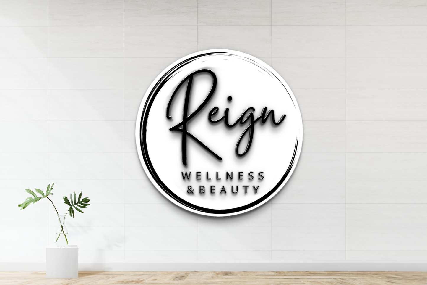 A large black-and-white circular 'Reign Wellness & Beauty' logo on a white tiled wall above a vase with green leaves.