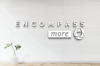 White tiled wall with 3D letters 'ENCOMPASS', script 'more' on white plaque, circular triple-loop logo, potted leaves.