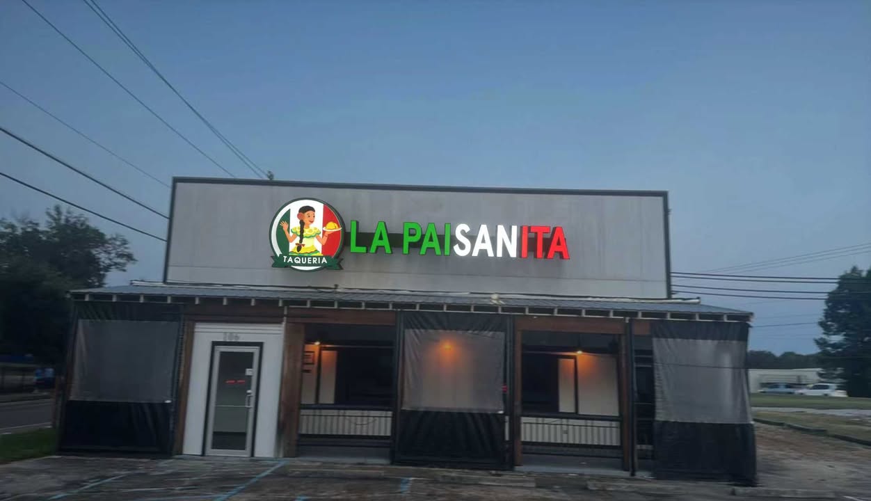 Gray storefront of La Paisanita Taqueria at dusk, illuminated green-white-red sign and round logo, covered patio.
