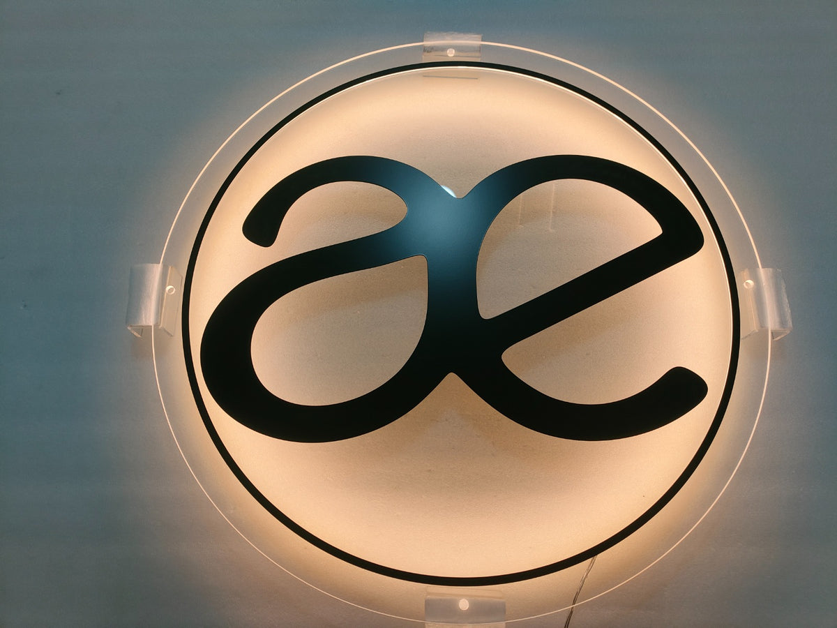 Wall-mounted round backlit sign with black stylized 'ae' logo on frosted warm light background, clear clips.