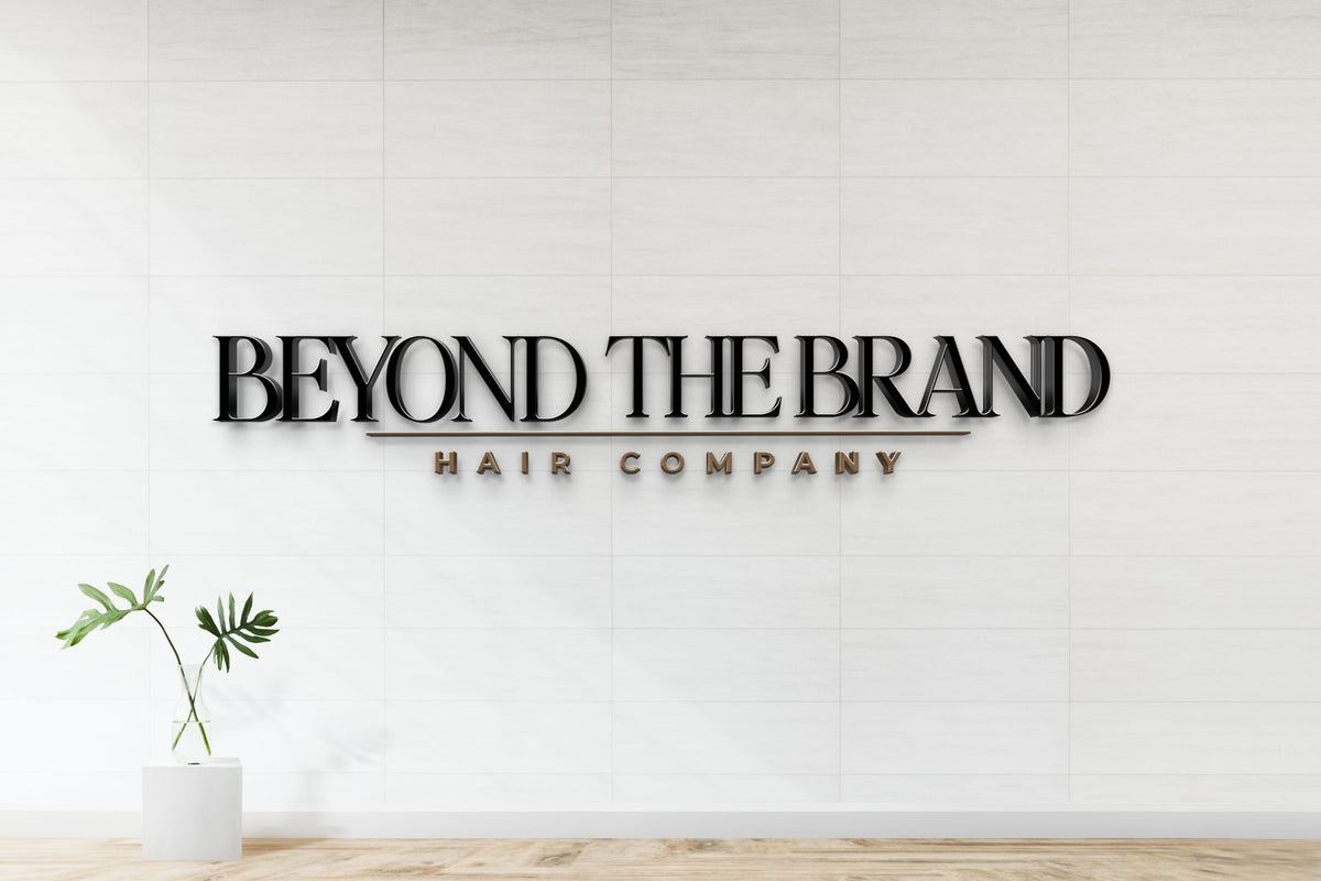 Black 3D BEYOND THE BRAND sign with gold underline and HAIR COMPANY on white tiled wall, glass vase with green leaves.