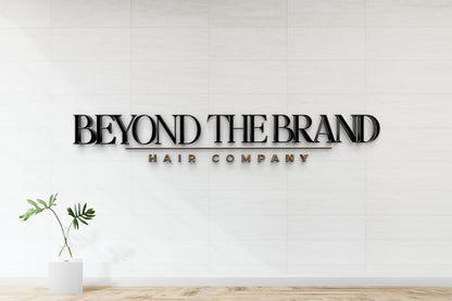 Black 3D BEYOND THE BRAND sign with gold underline and HAIR COMPANY on white tiled wall, glass vase with green leaves.