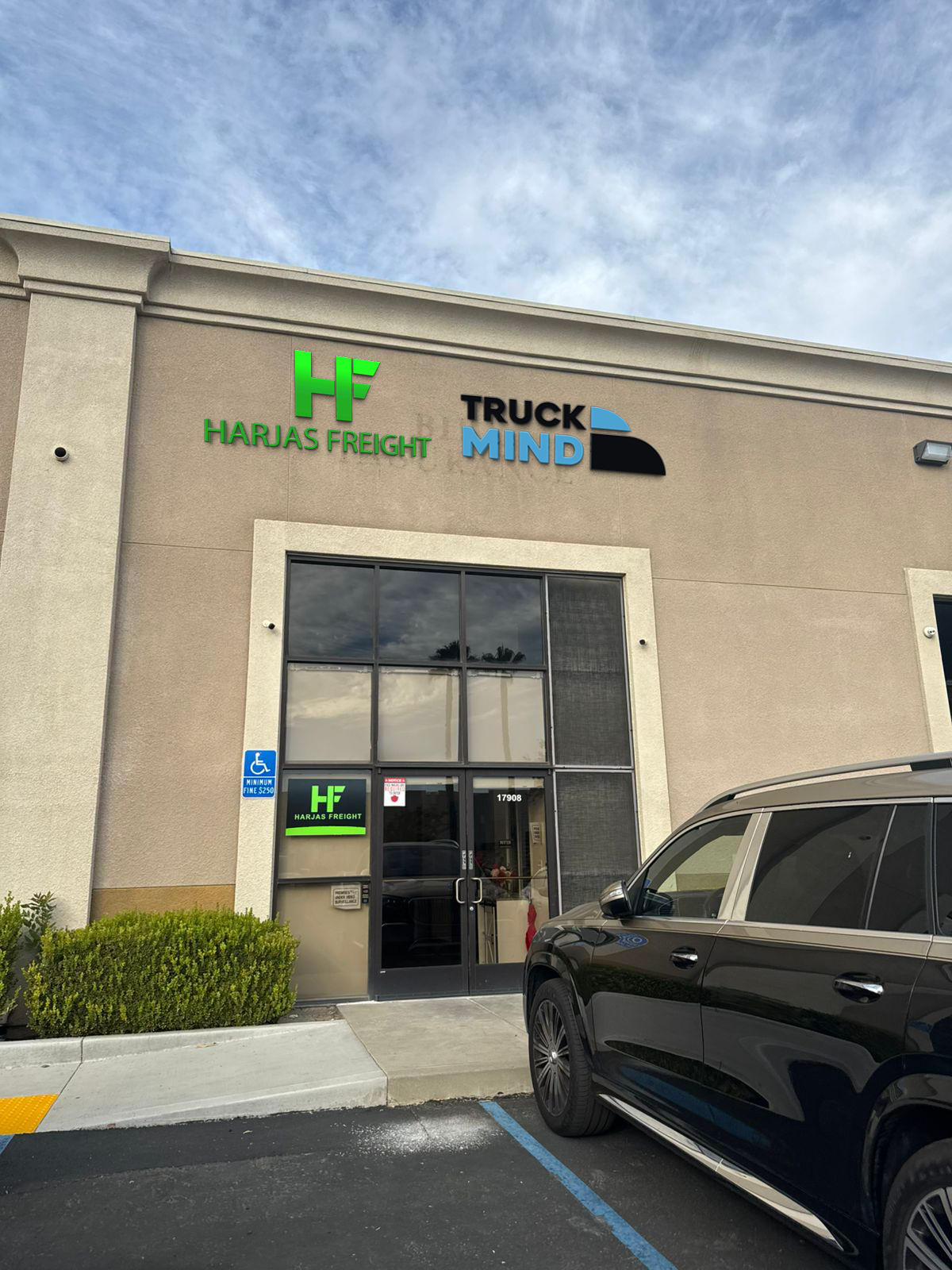 Tan stucco storefront with green HF Harjas Freight sign, blue-black TRUCK MIND logo, glass doors and black SUV.