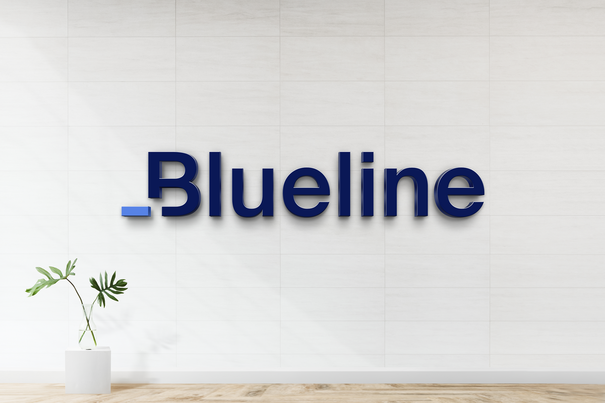 Dark blue 3D 'Blueline' wall sign with small blue dash on white tiled wall above wooden floor and potted plant.