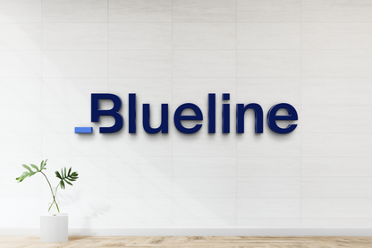 Dark blue 3D 'Blueline' wall sign with small blue dash on white tiled wall above wooden floor and potted plant.