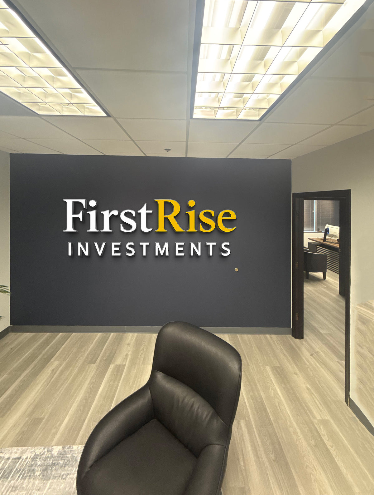 Office lobby with dark charcoal wall featuring FirstRise INVESTMENTS logo in white and yellow, black leather chair