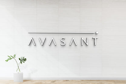 Metallic AVASANT logo and slim horizontal bar mounted on white tiled wall above glass vase with green leaves.