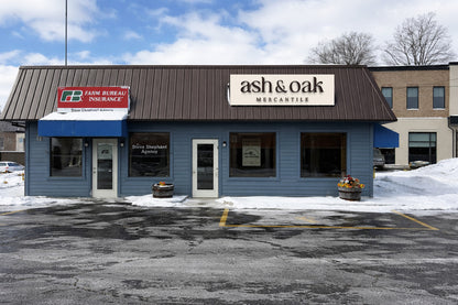 Blue-gray storefront with brown metal roof reading 'ash & oak mercantile', red Farm Bureau Insurance sign, snow.