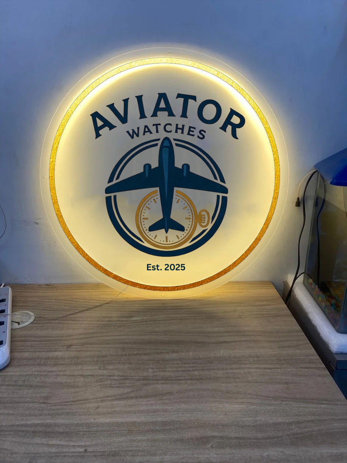 Illuminated round Aviator Watches sign with blue airplane and gold pocket-watch logo, gold rim, 'Est. 2025' on table.