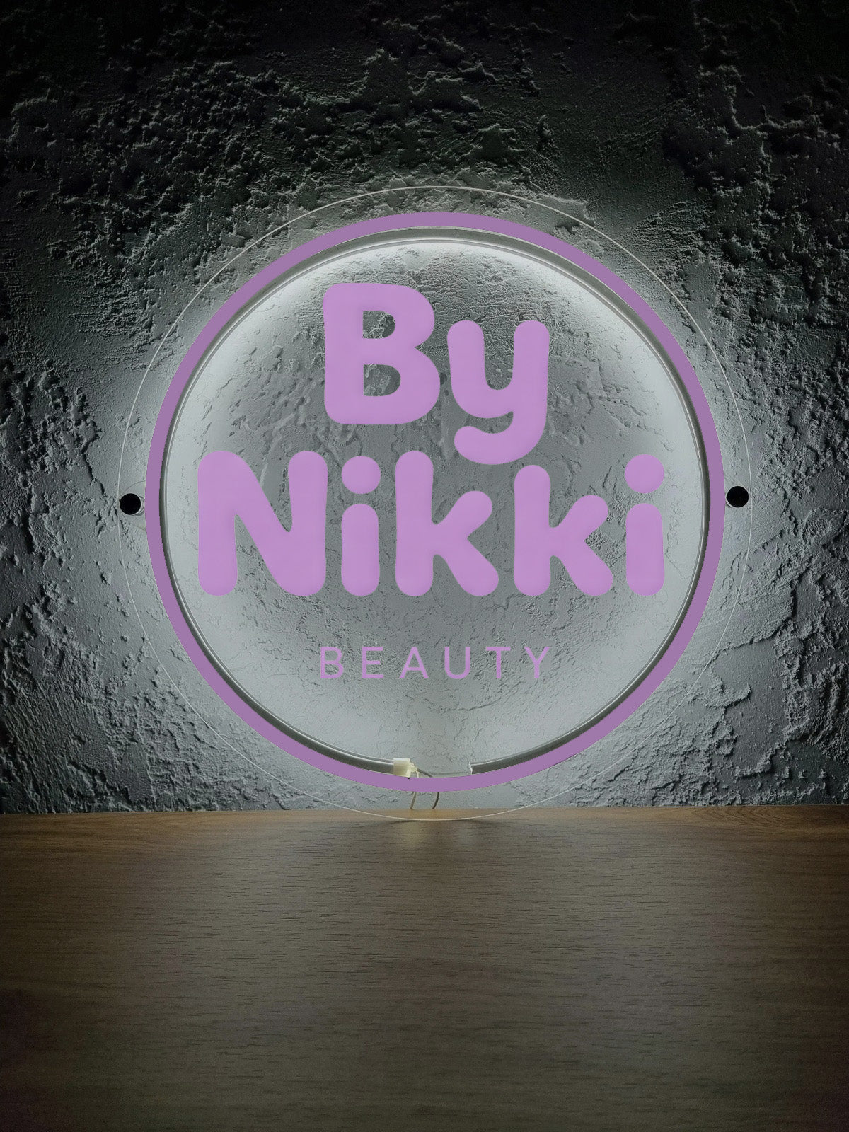 Illuminated round acrylic sign with purple 'By Nikki' and 'BEAUTY' text on textured gray wall above wooden counter.