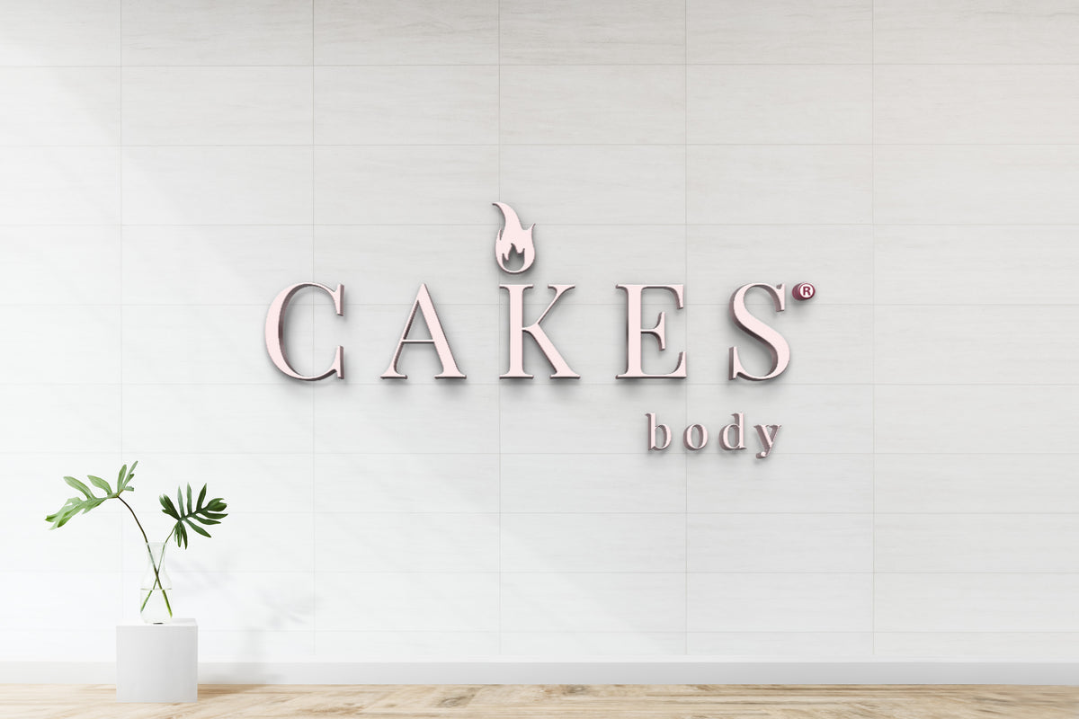 Pale pink metal CAKES sign with flame logo and small body text on white tiled wall; green plant in glass vase pedestal.