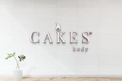 Pale pink metal CAKES sign with flame logo and small body text on white tiled wall; green plant in glass vase pedestal.