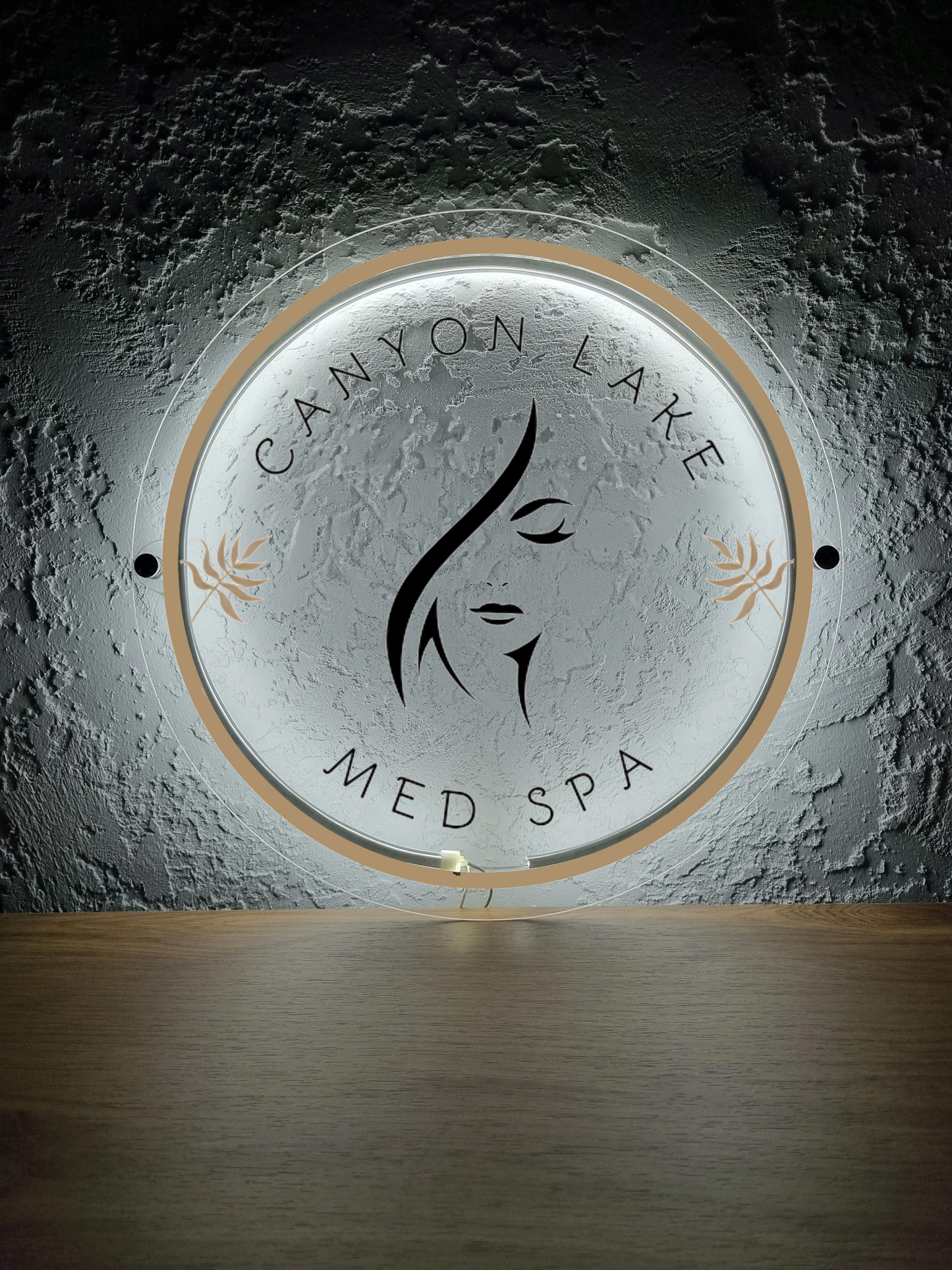 Illuminated round sign reading CANYON LAKE MED SPA with black stylized female face, gold accents on textured gray wall.