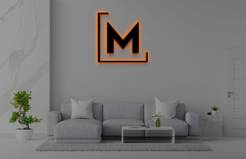 Black M logo with orange outline above light-gray sofa, marble wall panel, bonsai plant, coffee table with white.
