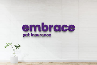 Purple 3D 'embrace' sign with smaller 'pet insurance' lettering on white tiled wall, vase with monstera leaves on stand.