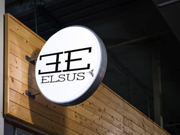 Round illuminated white sign with black stylized double-E logo and 'ELSUS' text mounted on wood plank wall indoors.