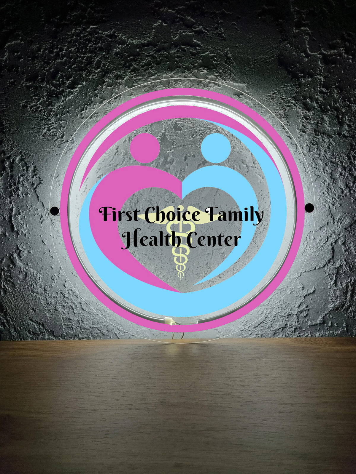 Circular neon logo First Choice Family Health Center with pink and blue hearts and yellow caduceus on textured gray.