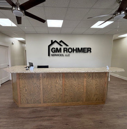 Office reception area with granite-topped wooden desk, wall logo "GM Rohmer Services, LLC", ceiling fans.