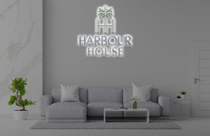 Modern gray living room with illuminated 'HARBOUR HOUSE' palm logo sign above sofa, coffee table and potted plants.