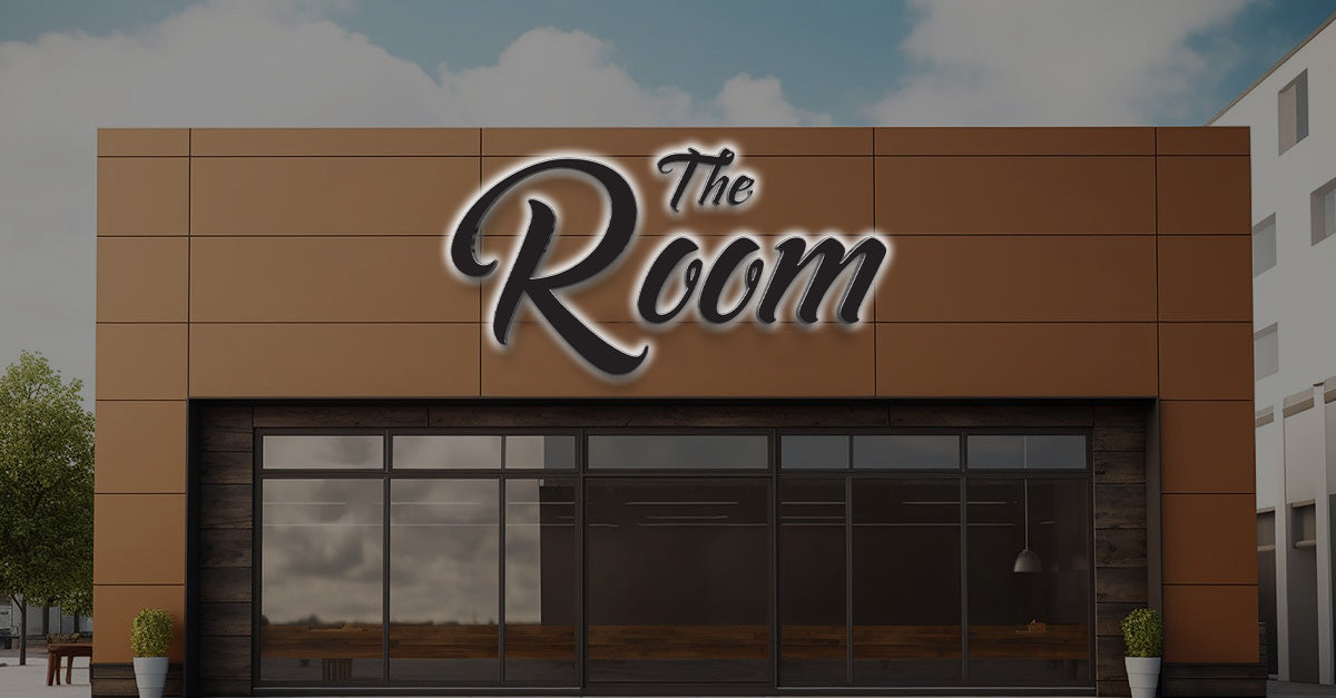 Brown panel storefront with large script sign The Room above dark glass windows, potted plants flanking entrance.