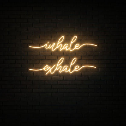 Glowing warm amber neon sign reading 'inhale' above 'exhale' in cursive, mounted on dark black brick wall.