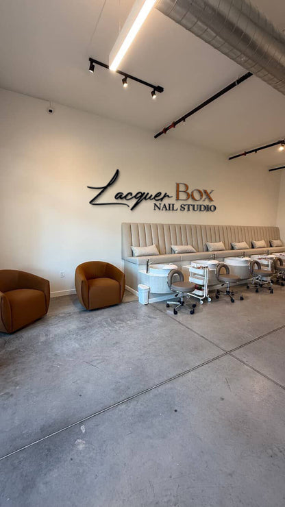 Wall sign 'Lacquer Box Nail Studio' above beige tufted bench, pedicure stations with rolling stools, brown armchairs.