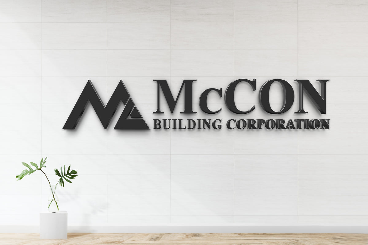 Black 3D 'McCON Building Corporation' wall sign with triangular M logo on white tiled wall and glass vase.