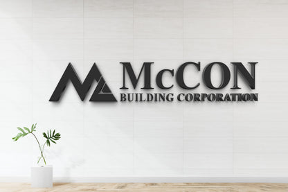 Black 3D 'McCON Building Corporation' wall sign with triangular M logo on white tiled wall and glass vase.