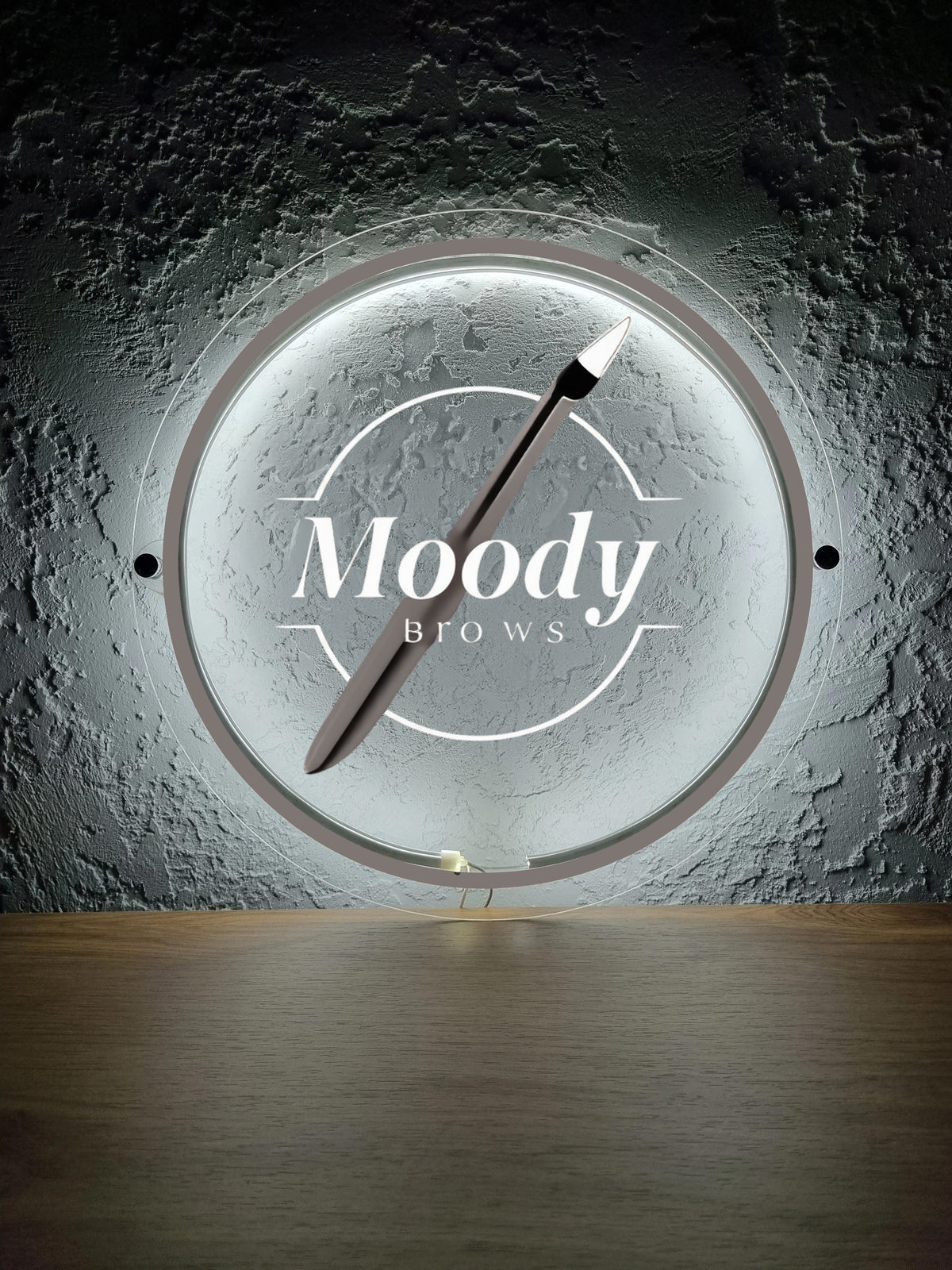 Illuminated round mirror with 'Moody Brows' logo, angled microblading pen across textured gray plaster wall.