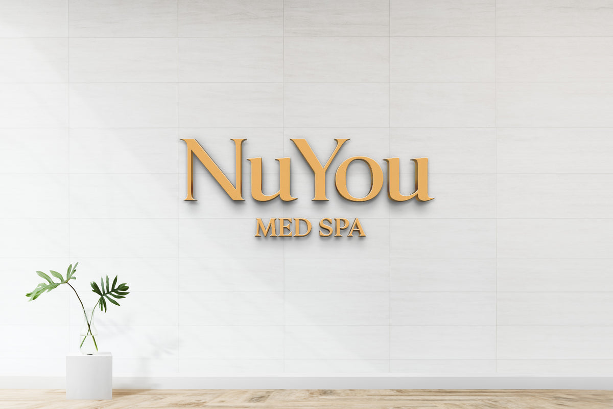Gold 3D NuYou sign with MED SPA lettering on white tiled wall, glass vase with green monstera on pedestal.