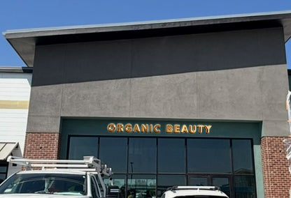 Storefront with gold 'ORGANIC BEAUTY' sign above dark glass windows, gray stucco facade, brick columns, white trucks.