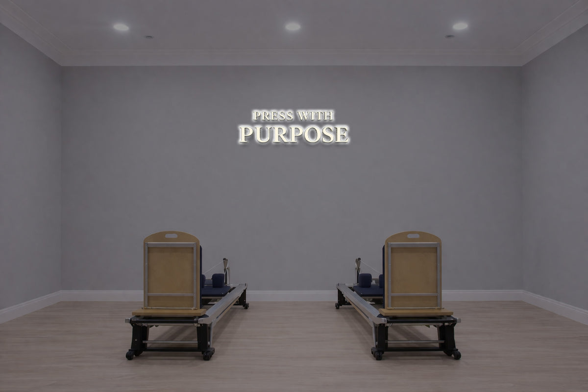 Minimalist gray Pilates studio with two wooden reformers on pale wood floor and illuminated PRESS WITH PURPOSE sign