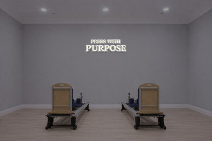 Minimalist gray Pilates studio with two wooden reformers on pale wood floor and illuminated PRESS WITH PURPOSE sign